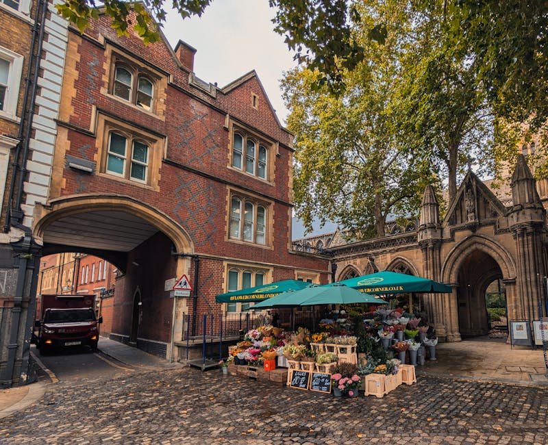 A bustling outdoor flower stall situated on a cobblestone street near Hoxton Overground station, operated by a floral florist. The stall is shaded by a green umbrella with the website 'flowerdeliveryh