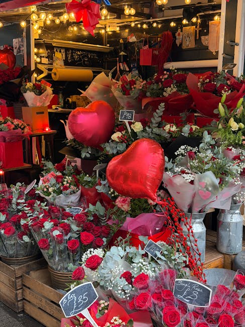 A vibrant display of fresh floral arrangements featuring red roses, white lilies, and sprigs of greenery, arranged in bouquets and wrapped in pink, red, and white tissue paper. Interspersed among the 